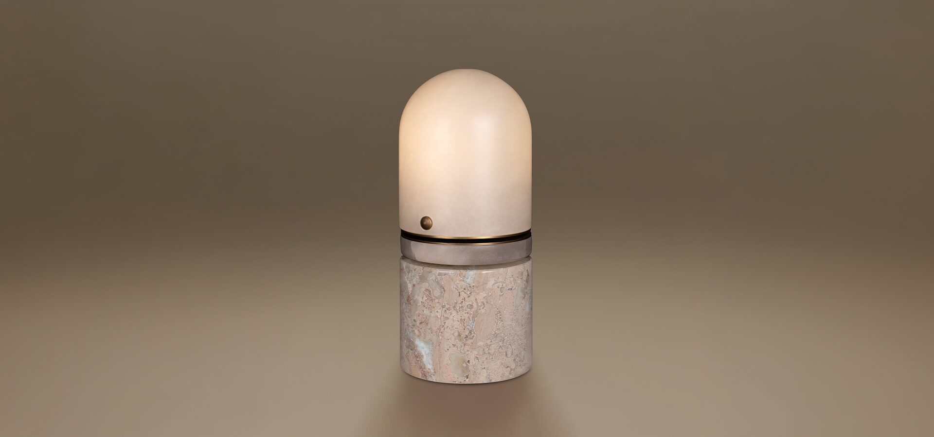 Russell Table Lamp by Porus Studio. A sophisticated, capsule-shaped luminaire featuring a rounded alabaster dome shade that provides a soft, warm diffusion of light. The design is structured with a central aged brushed brass ring detail, complete with a signature small metallic sphere. The lamp is supported by a solid, cylindrical base crafted from Breccia marble, showcasing its characteristic fragmented patterns and rich mineral textures. The piece is presented against a seamless, neutral-toned background.
