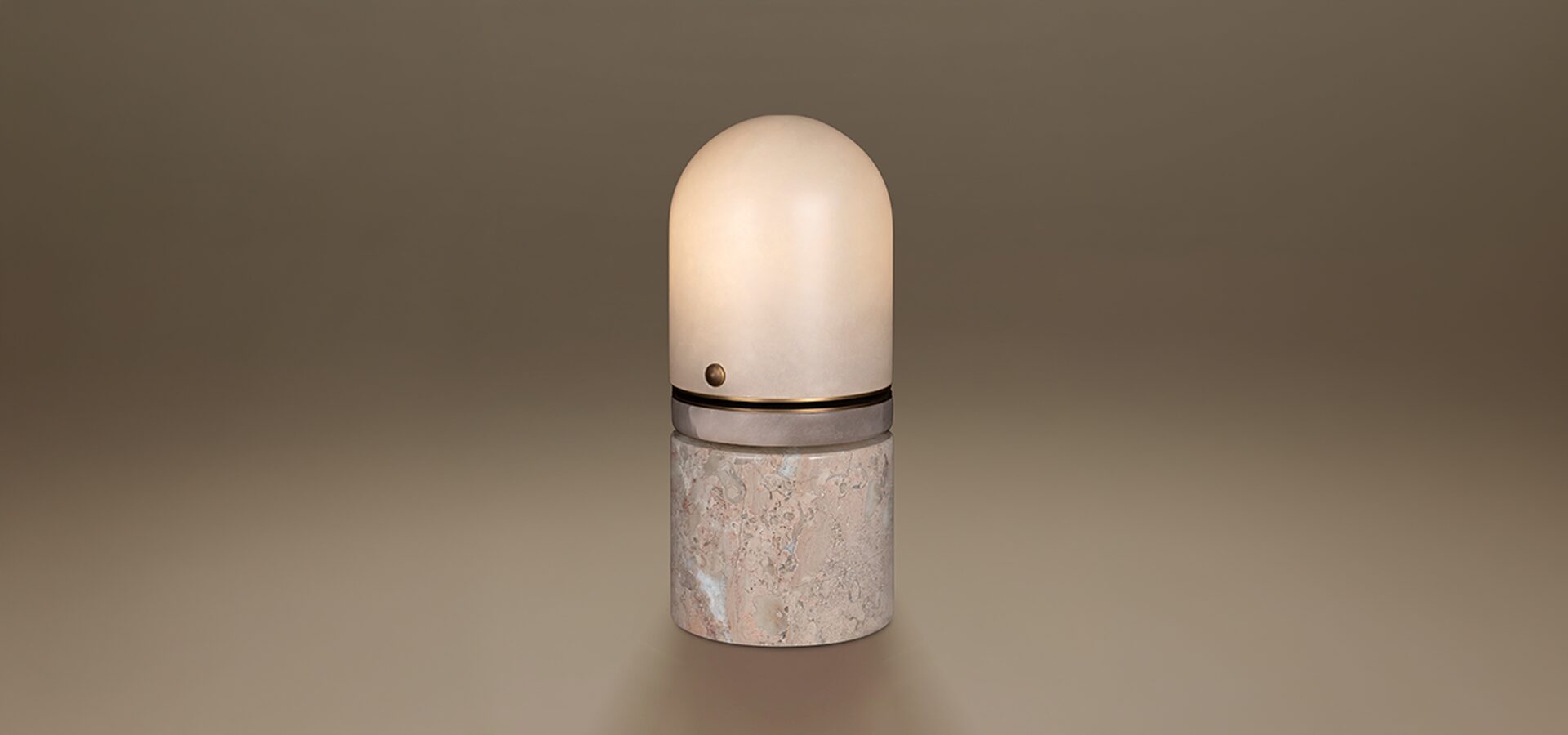 Russell Table Lamp by Porus Studio. A sophisticated, capsule-shaped luminaire featuring a rounded alabaster dome shade that provides a soft, warm diffusion of light. The design is structured with a central aged brushed brass ring detail, complete with a signature small metallic sphere. The lamp is supported by a solid, cylindrical base crafted from Breccia marble, showcasing its characteristic fragmented patterns and rich mineral textures. The piece is presented against a seamless, neutral-toned background.
