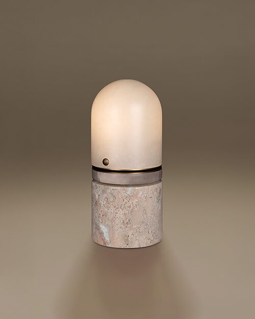 Russell Table Lamp by Porus Studio. A sophisticated, capsule-shaped luminaire featuring a rounded alabaster dome shade that provides a soft, warm diffusion of light. The design is structured with a central aged brushed brass ring detail, complete with a signature small metallic sphere. The lamp is supported by a solid, cylindrical base crafted from Breccia marble, showcasing its characteristic fragmented patterns and rich mineral textures. The piece is presented against a seamless, neutral-toned background.