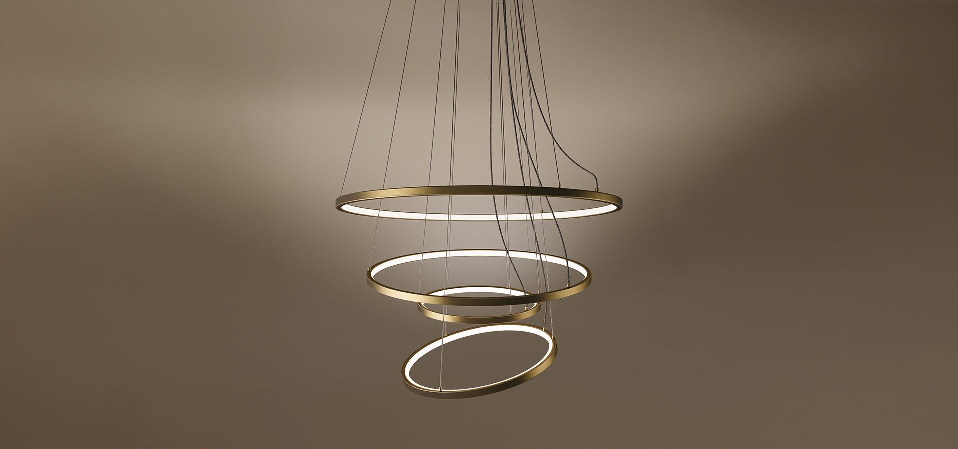 Gateway Suspension Lamp The Gateway suspension lamp, a modern multi-tier LED chandelier featuring four concentric rings with a sleek brass finish. Each ring is suspended by delicate cables, creating a dynamic, spiraling form. The interior of the rings emits a clean, warm light against a dark brown gradient background, showcasing a contemporary and architectural design.