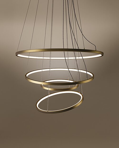 The Gateway suspension lamp, a modern multi-tier LED chandelier featuring four concentric rings with a sleek brass finish. Each ring is suspended by delicate cables, creating a dynamic, spiraling form. The interior of the rings emits a clean, warm light against a dark brown gradient background, showcasing a contemporary and architectural design.