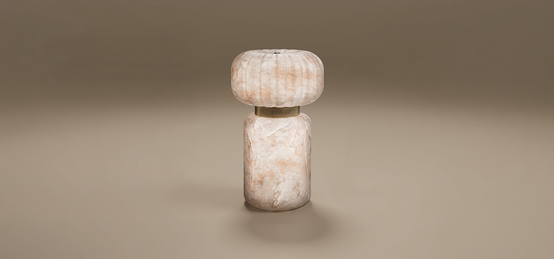 Hampshire Table Lamp Hampshire Table Lamp by Porus Studio. A contemporary sculptural lamp crafted entirely from translucent alabaster with natural peach and cream tones. The design features a heavy, cylindrical pedestal base and a rounded, ribbed top shade. Connecting the two stone elements is a aged brushed brass ring engraved with the Porus Studio logo. The lamp glows softly from within, emphasizing the organic patterns of the stone.