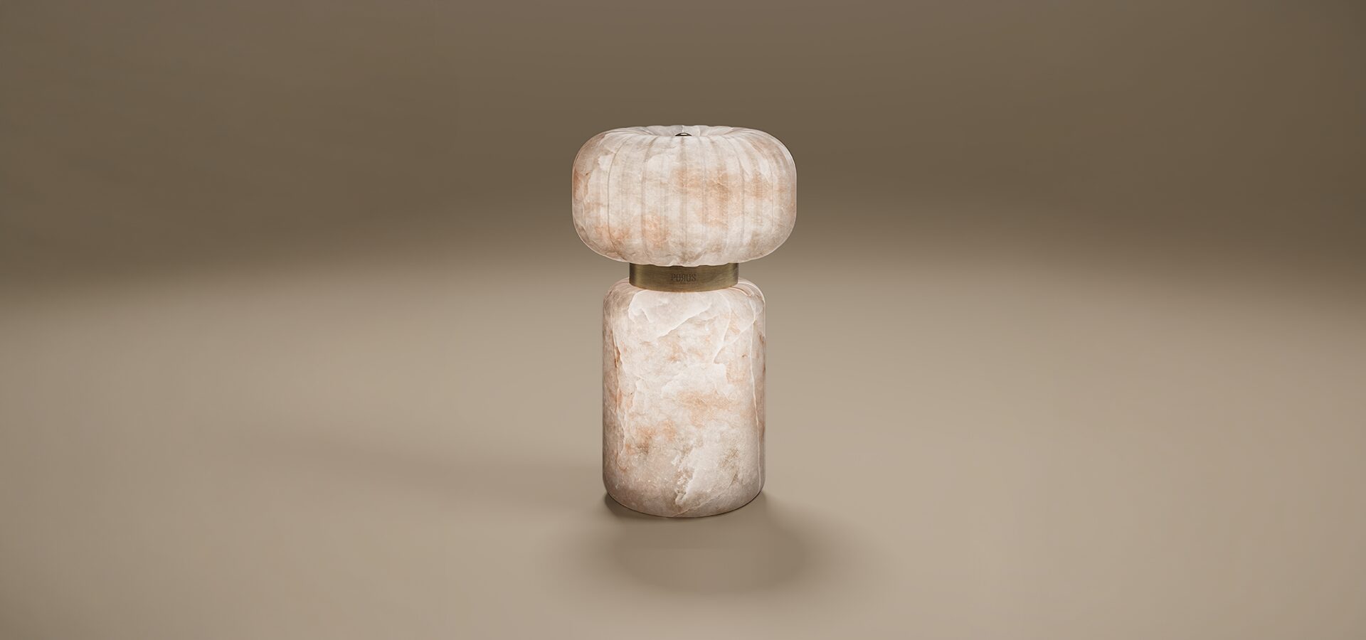 Hampshire Table Lamp by Porus Studio. A contemporary sculptural lamp crafted entirely from translucent alabaster with natural peach and cream tones. The design features a heavy, cylindrical pedestal base and a rounded, ribbed top shade. Connecting the two stone elements is a aged brushed brass ring engraved with the Porus Studio logo. The lamp glows softly from within, emphasizing the organic patterns of the stone.