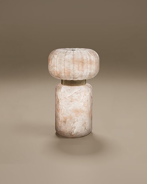 Hampshire Table Lamp by Porus Studio. A contemporary sculptural lamp crafted entirely from translucent alabaster with natural peach and cream tones. The design features a heavy, cylindrical pedestal base and a rounded, ribbed top shade. Connecting the two stone elements is a aged brushed brass ring engraved with the Porus Studio logo. The lamp glows softly from within, emphasizing the organic patterns of the stone.