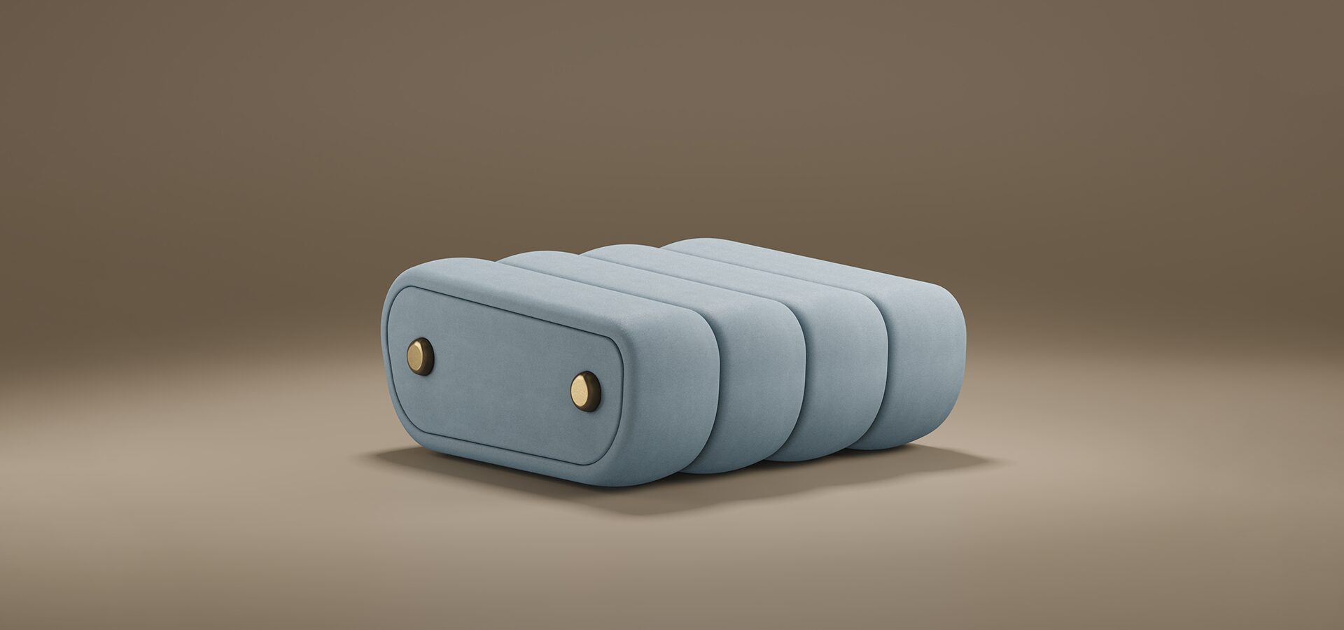 A product image of the Dakota Ottoman by Porus Studio against a muted brown background. The ottoman has a unique, low-profile silhouette composed of four thick, rounded vertical segments upholstered in a smooth, light blue or slate grey velvet. The side panel is accented with two circular brass metal buttons, giving it a luxurious, tailored appearance reminiscent of vintage luggage or high-end mid-century furniture.