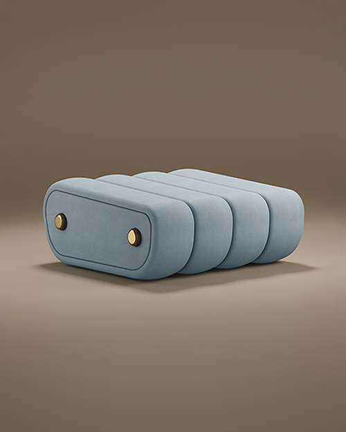 A product image of the Dakota Ottoman by Porus Studio against a muted brown background. The ottoman has a unique, low-profile silhouette composed of four thick, rounded vertical segments upholstered in a smooth, light blue or slate grey velvet. The side panel is accented with two circular brass metal buttons, giving it a luxurious, tailored appearance reminiscent of vintage luggage or high-end mid-century furniture.