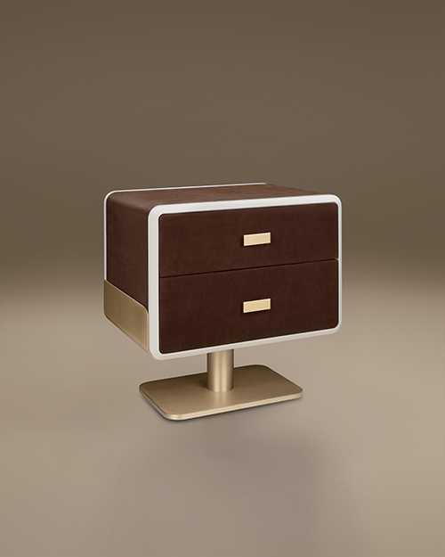 A luxury Phoenix Bedside Table by Porus Studio, featuring a retro-inspired, rounded rectangular body upholstered in dark brown leather. The face of the two drawers is framed by a lacquered trim that provides a sharp metallic contrast. The unit is supported by a central pedestal base with minimalist horizontal handles, both finished in brushed brass. The design blends Space Age aesthetics with premium materials, set against a neutral, warm studio background.