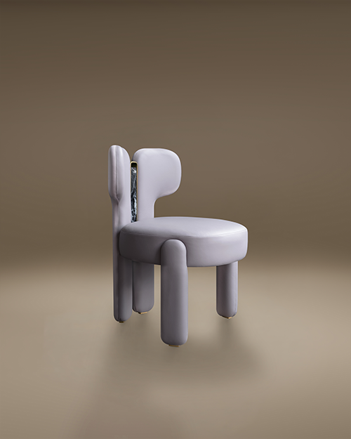 A modern dining chair with a bold, avant-garde design. The Idaho Dining Chair by Porus Studio features a plush, rounded seat and backrest upholstered in a soft, leather. Its thick, cylindrical legs and asymmetrical backrest give it a sculptural, "chubby" aesthetic. A striking vertical insert of dark, veined marble framed in polished brass accents the backrest, adding a touch of luxury to the contemporary, playful silhouette. The chair is set against a neutral, minimalist taupe background.