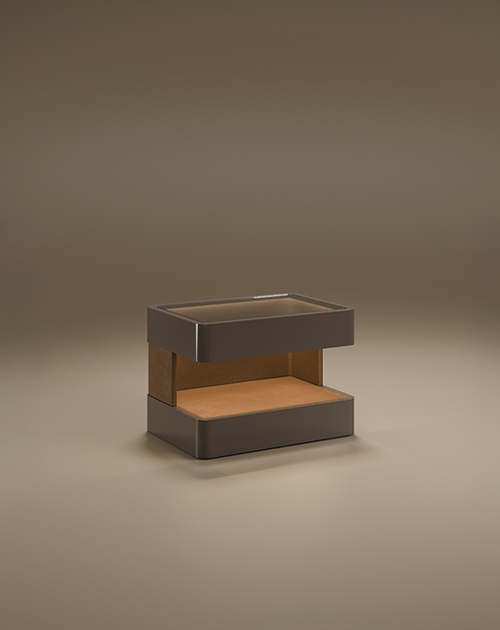 A luxury Creek I Side Table by Porus Studio, featuring a rectangular design with rounded edges and a large horizontal opening. The outer structure is finished in a dark grey high-gloss lacquer, while the interior display nook is lined with warm tan leather. The top surface includes a smoke glass inlay. The piece is shown in a studio setting against a neutral, warm-toned background.