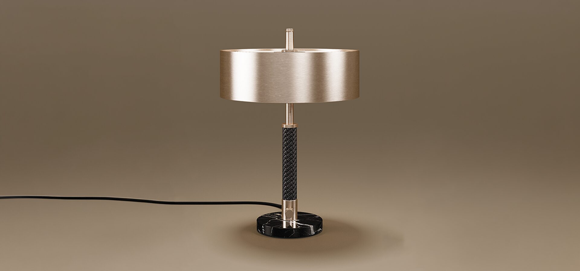 A sophisticated Chicago Table Lamp set against a neutral taupe background. The lamp features a wide, cylindrical drum shade in a brushed brass finish. The central stem is detailed with a textured black braided leather wrap, bookended by polished brass accents. The fixture sits on a solid, circular base made of black marble with white veining. A black power cord extends from the side of the base.