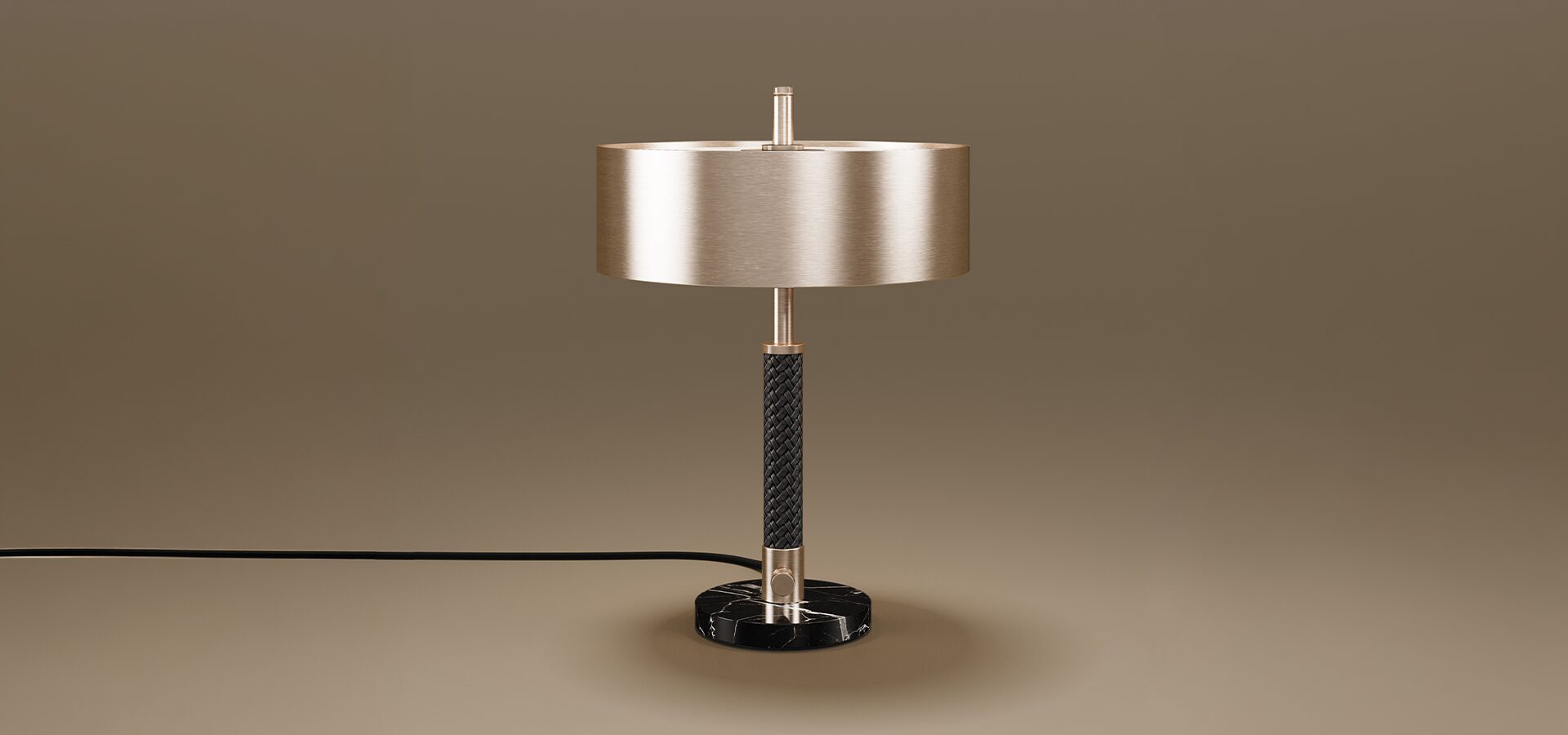 A sophisticated Chicago Table Lamp set against a neutral taupe background. The lamp features a wide, cylindrical drum shade in a brushed brass finish. The central stem is detailed with a textured black braided leather wrap, bookended by polished brass accents. The fixture sits on a solid, circular base made of black marble with white veining. A black power cord extends from the side of the base.