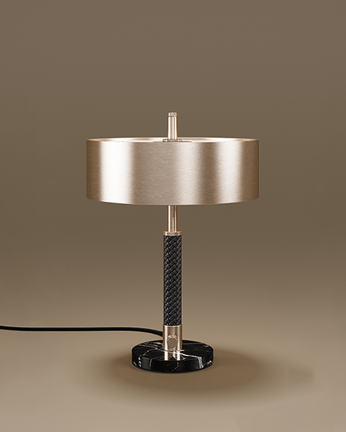 A sophisticated Chicago Table Lamp set against a neutral taupe background. The lamp features a wide, cylindrical drum shade in a brushed brass finish. The central stem is detailed with a textured black braided leather wrap, bookended by polished brass accents. The fixture sits on a solid, circular base made of black marble with white veining. A black power cord extends from the side of the base.