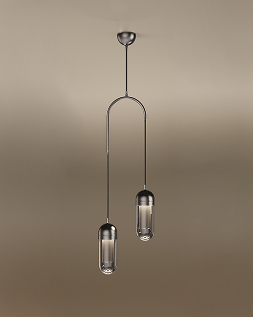 The Denver II pendant lamp by Porus Studio, featuring two sophisticated capsule-shaped light fixtures suspended at different heights from a dark metallic arch. Each fixture has a dark metal cap and a transparent glass diffuser enclosing a glowing internal mesh cylinder. A single central cable connects the arch to a dark metal ceiling canopy. The entire lamp is set against a soft, warm-toned studio background.