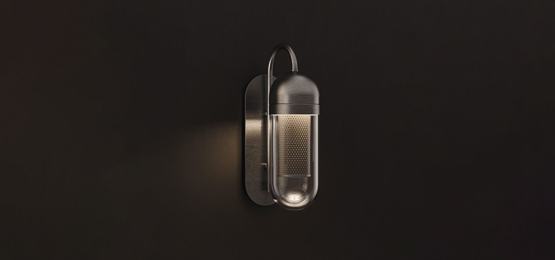 Denver Wall Lamp Denver Wall Lamp, featuring a more industrial brushed metal or aged brass finish. The lamp is illuminated against a pitch-black background, creating a dramatic chiaroscuro effect. The warm light filters through the perforated stainless steel core and reflects off the clear glass outer shell, casting a focused glow back onto the metallic mounting plate.