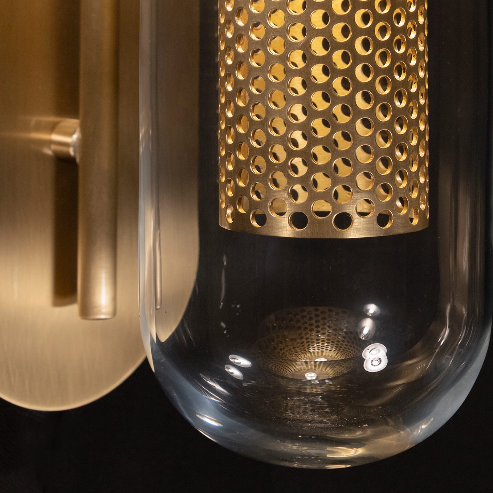 Denver Wall Lamp Close-up detail of the lower part of the Denver Wall Lamp against a black background. The image showcases a rounded, transparent clear glass capsule enclosing an inner perforated cylinder made of gold brushed stainless steel. On the left, a portion of the matching gold brushed metal wall mount and vertical rod is visible.