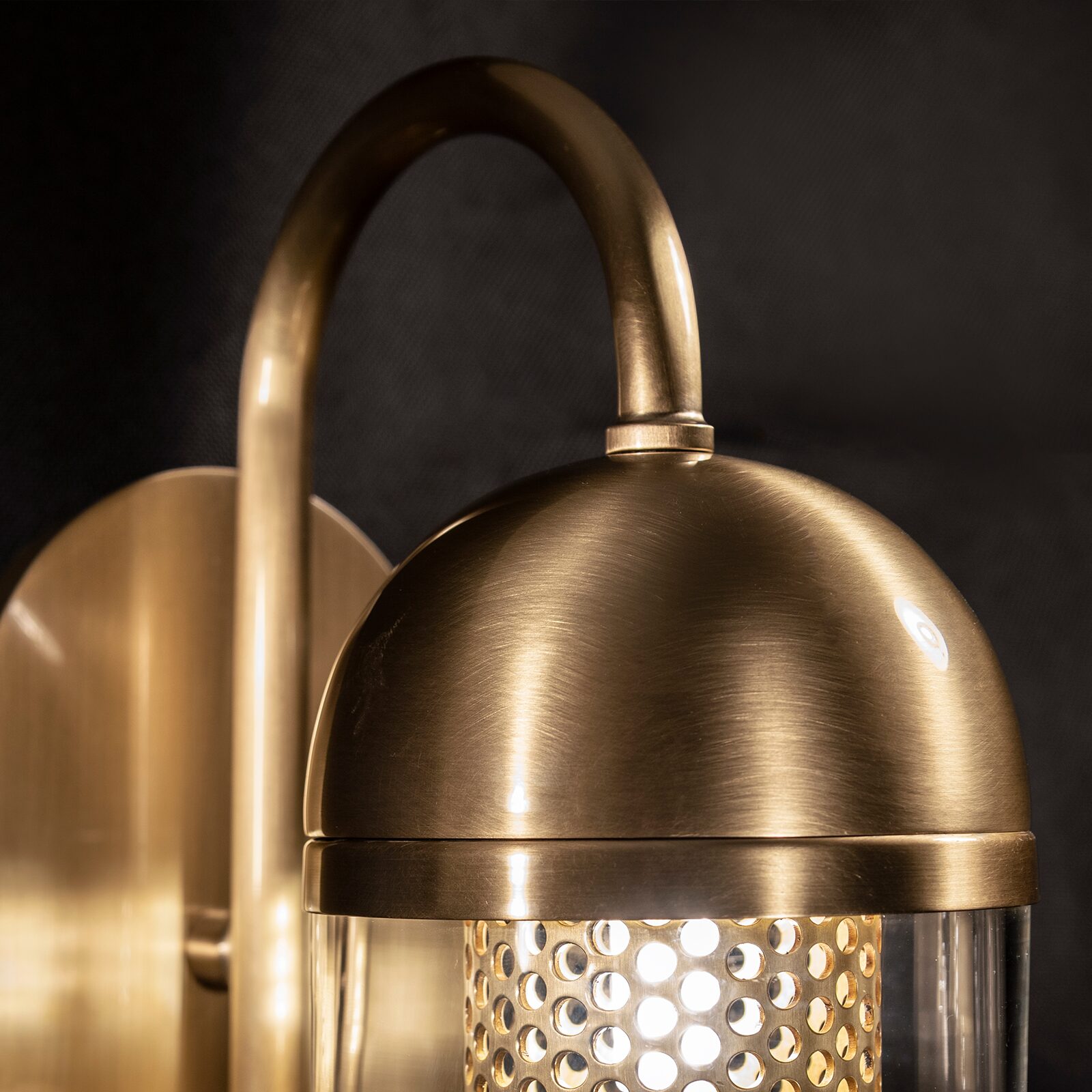Denver Wall Lamp Close-up detail of the top part of the Denver Wall Lamp against a dark backdrop. The image features a dome-shaped cap and a curved gooseneck arm, both crafted from textured gold brushed stainless steel. Below the dome, the illuminated light source is partially visible through a perforated inner metal mesh and clear glass capsule.