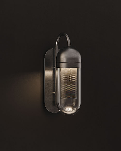 A vertical thumbnail image of the Denver Wall Lamp, featuring a more industrial brushed metal or aged brass finish. The lamp is illuminated against a pitch-black background, creating a dramatic chiaroscuro effect. The warm light filters through the perforated stainless steel core and reflects off the clear glass outer shell, casting a focused glow back onto the metallic mounting plate.