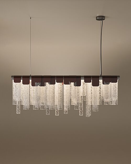 The Frostlight suspension lamp, a sophisticated linear lamp featuring a row of textured, frosted glass panels in varying lengths. The glass slabs are held by elegant dark leather or metal clasps along a horizontal bar, creating a rhythmic, shimmering effect that resembles hanging ice or frost. The fixture is suspended by two thin cables against a warm, neutral background, blending contemporary design with organic textures.