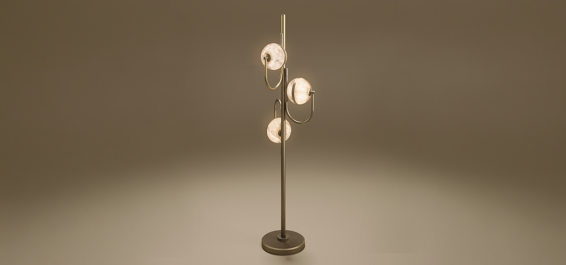 Oculus floor lamp in aged brushed brass, lit, with three glowing alabaster discs staggered along the vertical stem, casting a warm pool of light on a taupe gradient background.