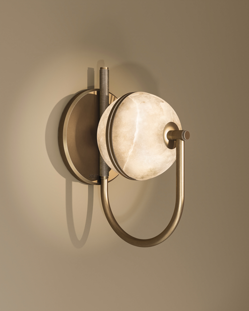 Oculus wall lamp in aged brushed brass with a glowing alabaster disc, mounted on a round backplate and held by a U-shaped tubular arm, casting a soft shadow on a warm beige wall.